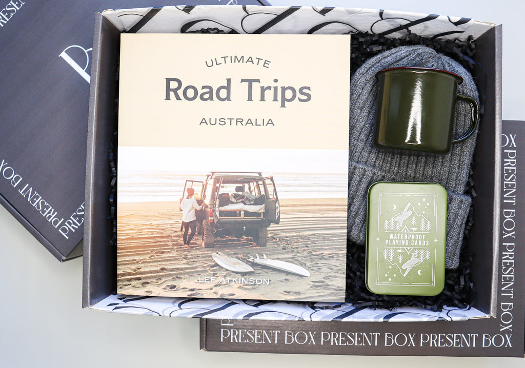 Charcoal box filled with Ultimate Road Trips Australia book, grey beanie, black camping mug and green metal tin of playing card