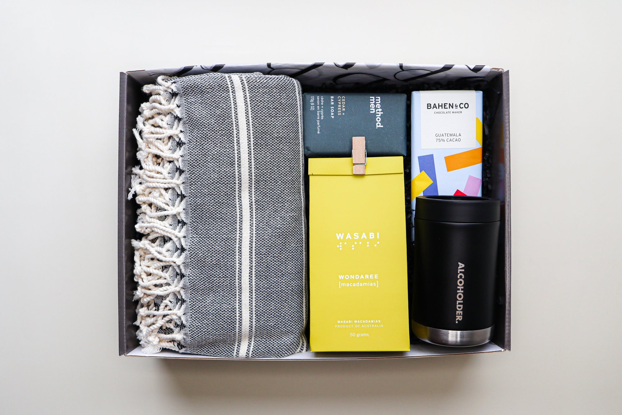 A beautiful gift box containing a grey and white striped Turkish Towel, a Method Man Bar Soap, a yellow pack of Wondaree Wasabi Macadamias closed a the top with a timber peg, a colourful bar of Bahen & Co Chocolate and a black and steel Alcoholder