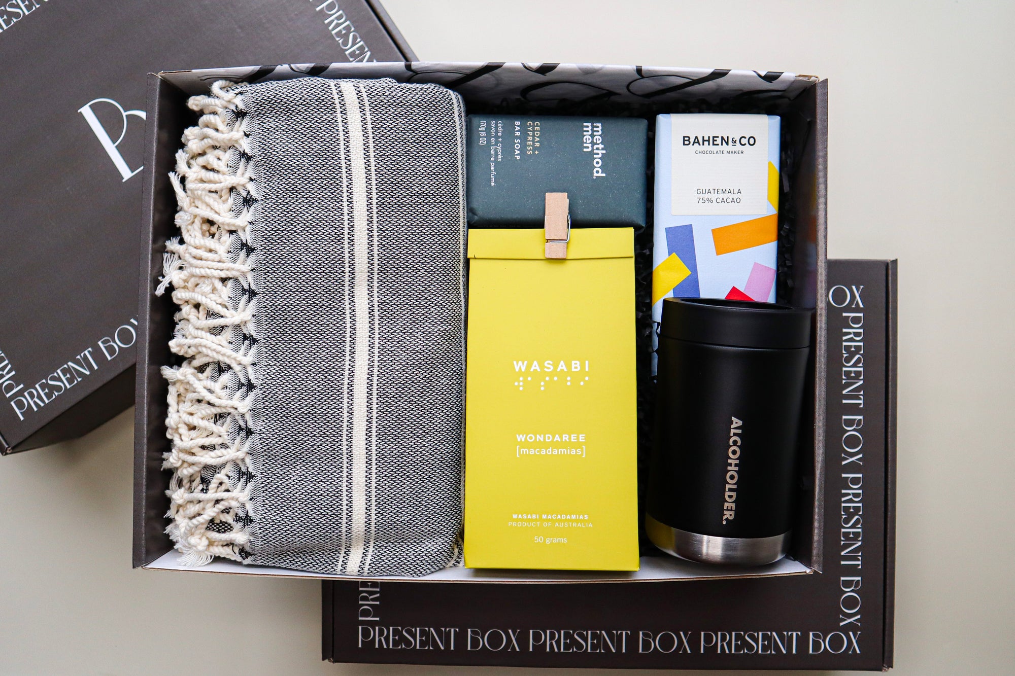 A beautiful gift box containing a grey and white striped Turkish Towel, a Method Man Bar Soap, a yellow pack of Wondaree Wasabi Macadamias closed a the top with a timber peg, a colourful bar of Bahen & Co Chocolate and a black and steel Alcoholder