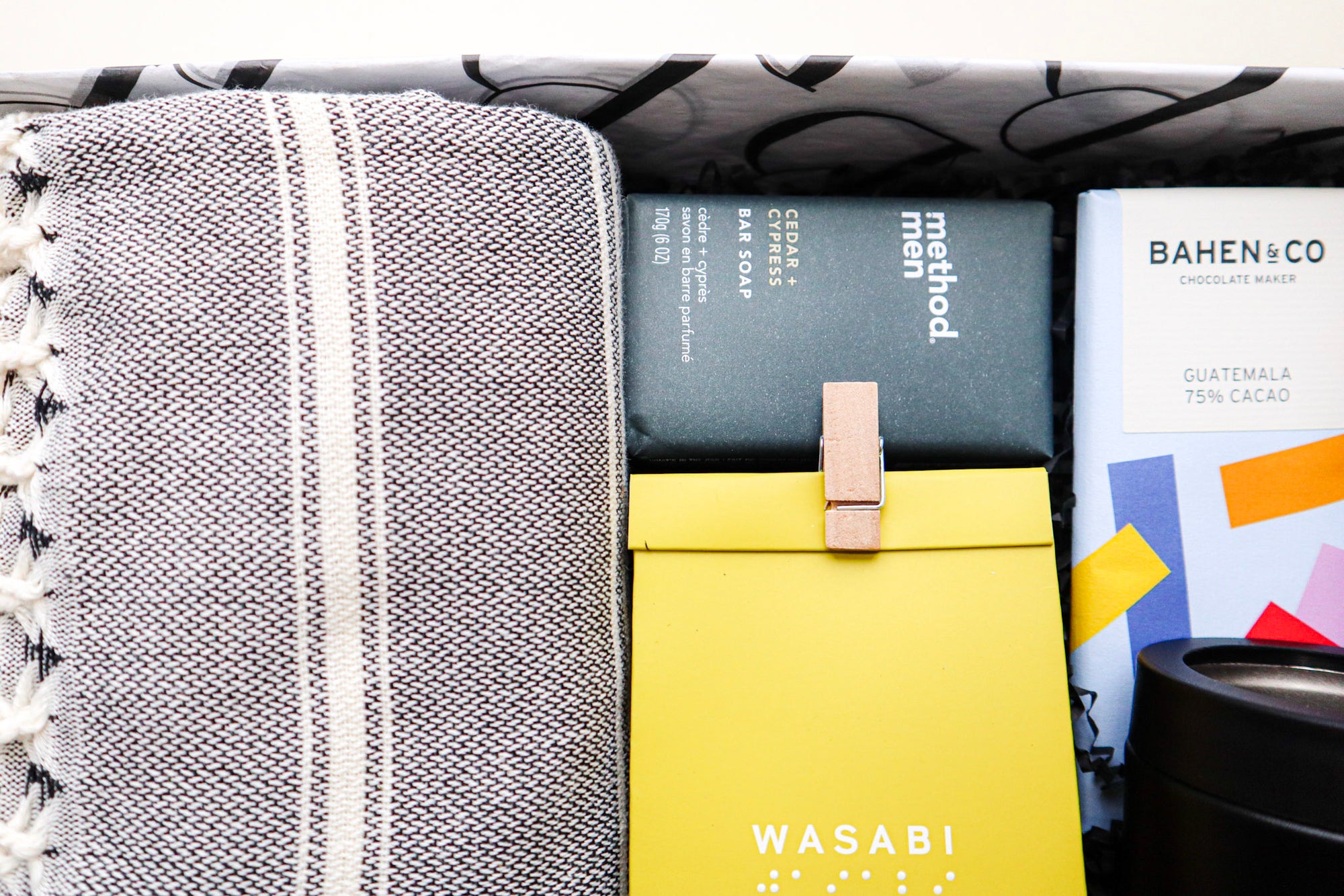 A beautiful gift box containing a grey and white striped Turkish Towel, a Method Man Bar Soap, a yellow pack of Wondaree Wasabi Macadamias closed a the top with a timber peg, a colourful bar of Bahen & Co Chocolate and a black and steel Alcoholder