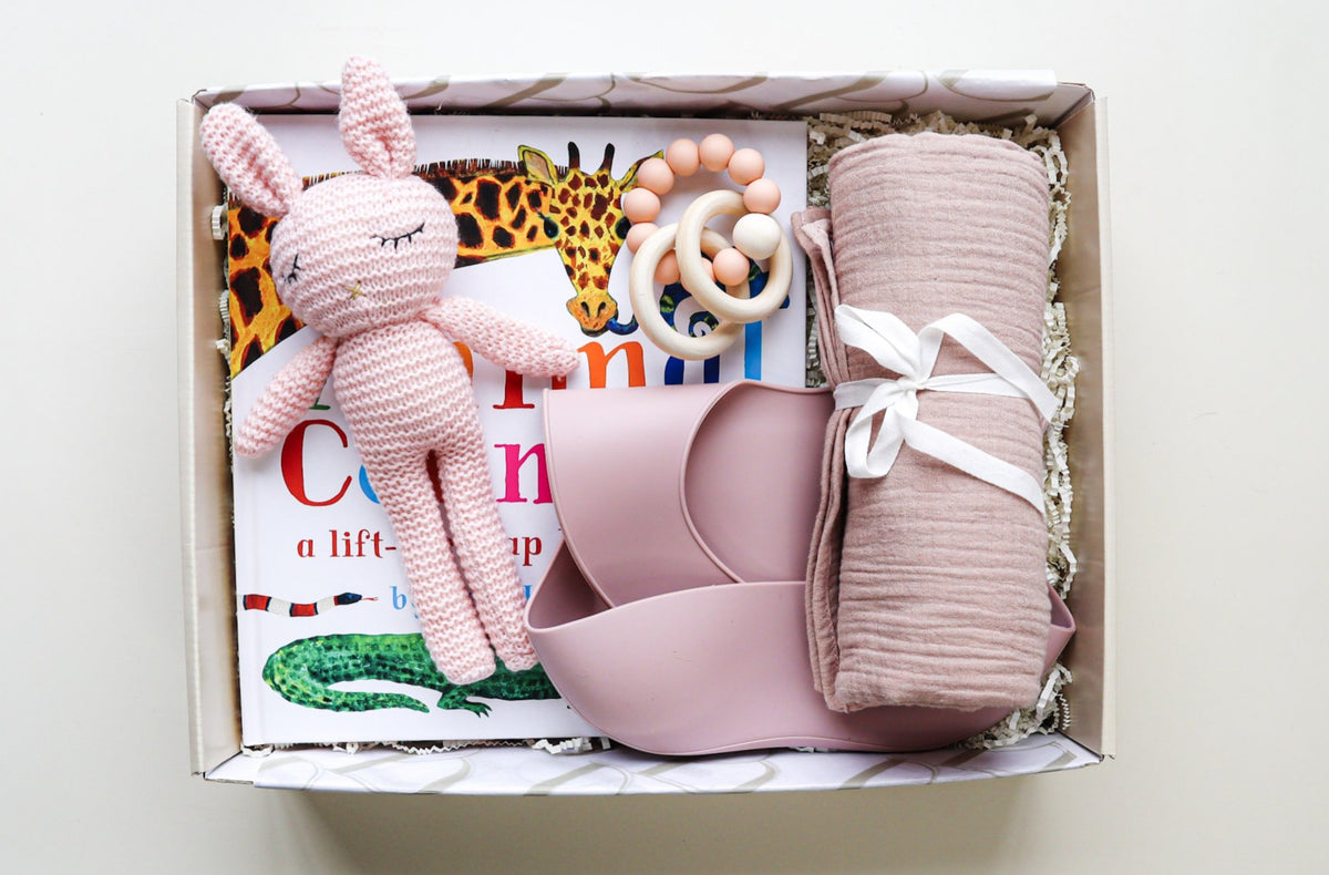 It's A Girl Gift Box is a cream cardboard box containing a pink crocheted bunny, a pink silicone baby bib, a pink cotton wrap rolled and tied with a white ribbon, a pink and natural coloured teether ring and rattle toy and a children's hardcover book titled Animal Counting