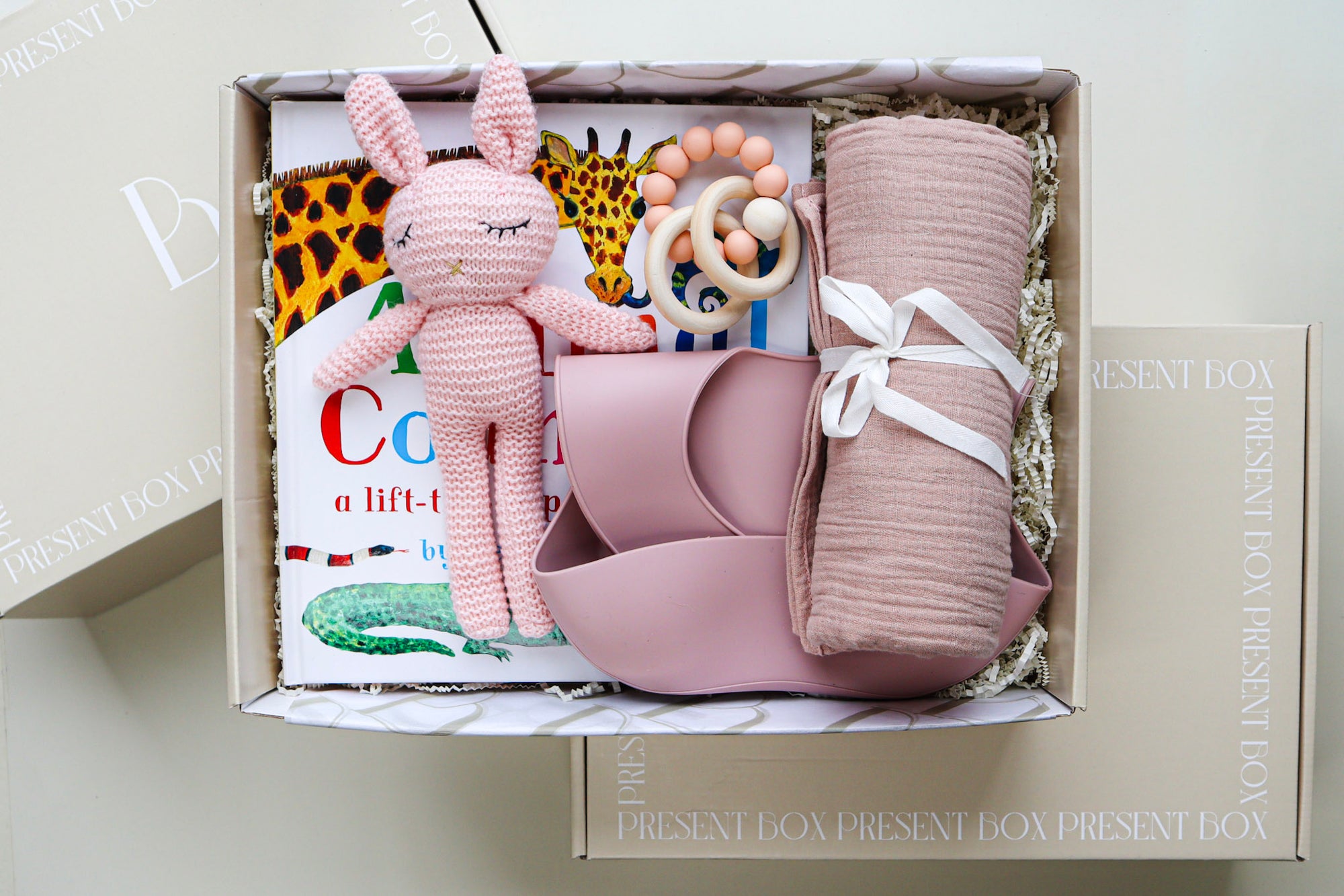 It's A Girl Gift Box is a cream cardboard box containing a pink crocheted bunny toy, a pink silicone baby bib, a pink cotton wrap rolled and tied with a white ribbon, a pink and natural coloured teether ring and rattle toy and a children's hardcover book titled Animal Counting