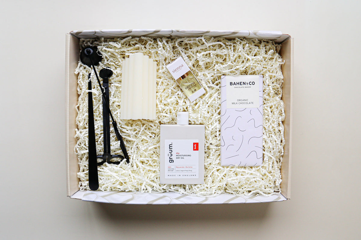 A beautiful natural coloured gift box containing a 3 piece matt black metal candle care set, a cream pillar candle, white packaged Sienna Cuticle Oil, a white bottle of Gruum Moisturising Dry Oil and a black and white bar of Bahen & Co Organic Milk Chocolate all nestled amongst  natural paper raffia