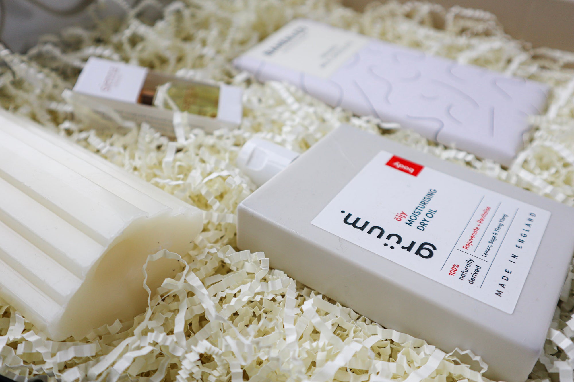 A white bottle of Gruum Moisturising Dry Oil and a black and white bar of Bahen & Co Organic Milk Chocolate all nestled amongst natural paper raffia