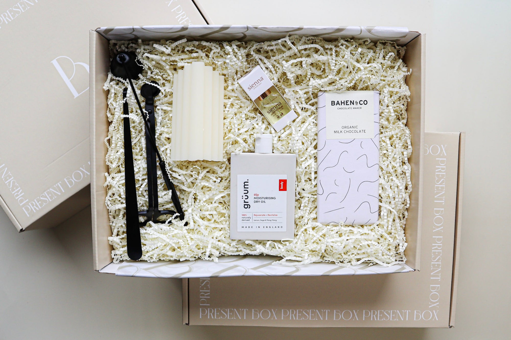 A beautiful natural coloured gift box containing a 3 piece matt black metal candle care set, a cream pillar candle, white packaged Sienna Cuticle Oil, a white bottle of Gruum Moisturising Dry Oil and a black and white bar of Bahen & Co Organic Milk Chocolate all nestled amongst  natural paper raffia