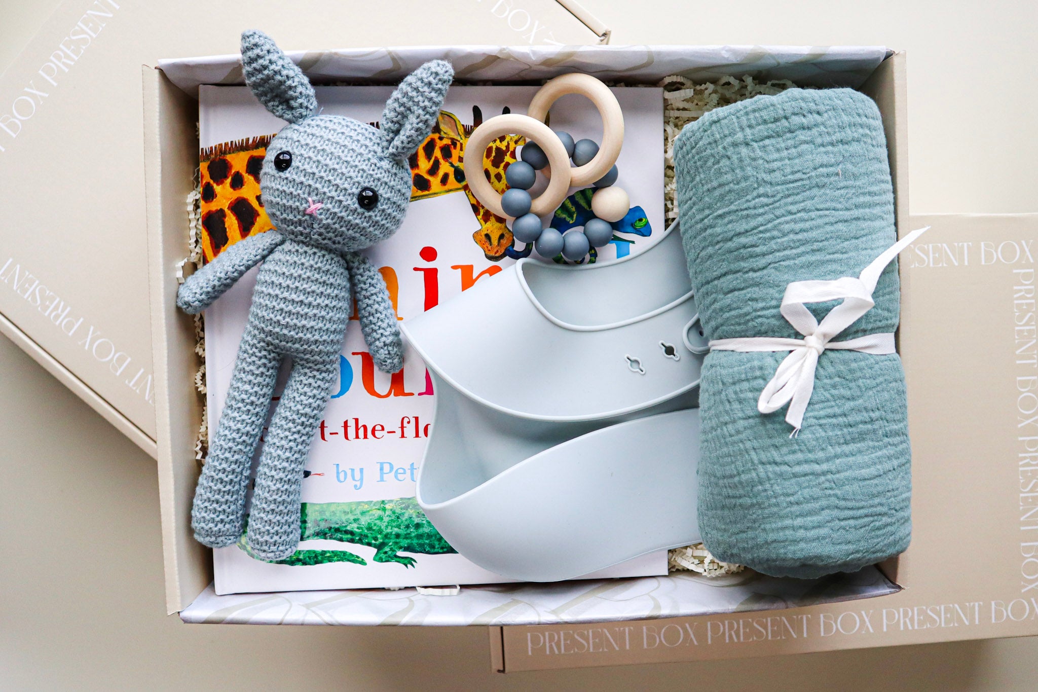 It's A Boy Gift Box is a cream cardboard box containing a blue, crocheted bunny, a light blue silicone bib, a blue cotton wrap rolled and tied with a white ribbon, a blue and natural coloured teether ring and rattle toy and a children's hardcover book titled Animal Counting