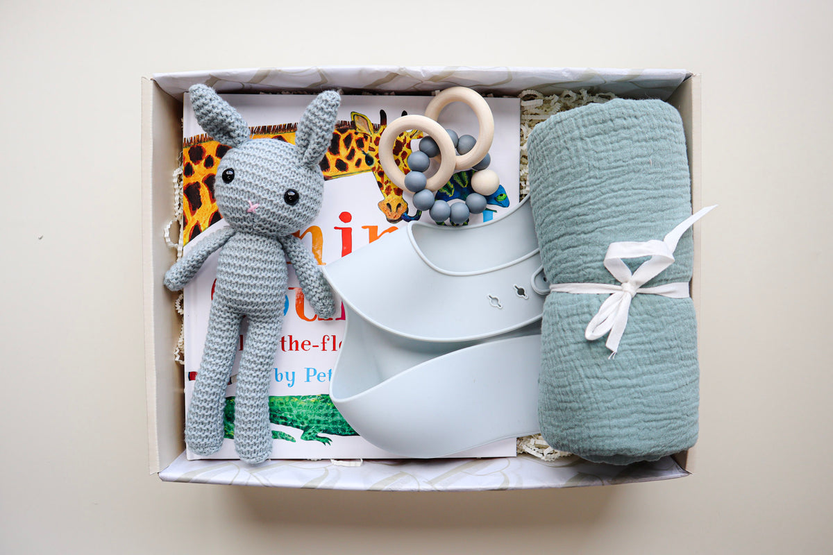 It's A Boy Gift Box is a cream cardboard box containing a blue, crocheted bunny, a light blue silicone bib, a blue cotton wrap rolled and tied with a white ribbon, a blue and natural coloured teether ring and rattle toy and a children's hardcover book titled Animal Counting