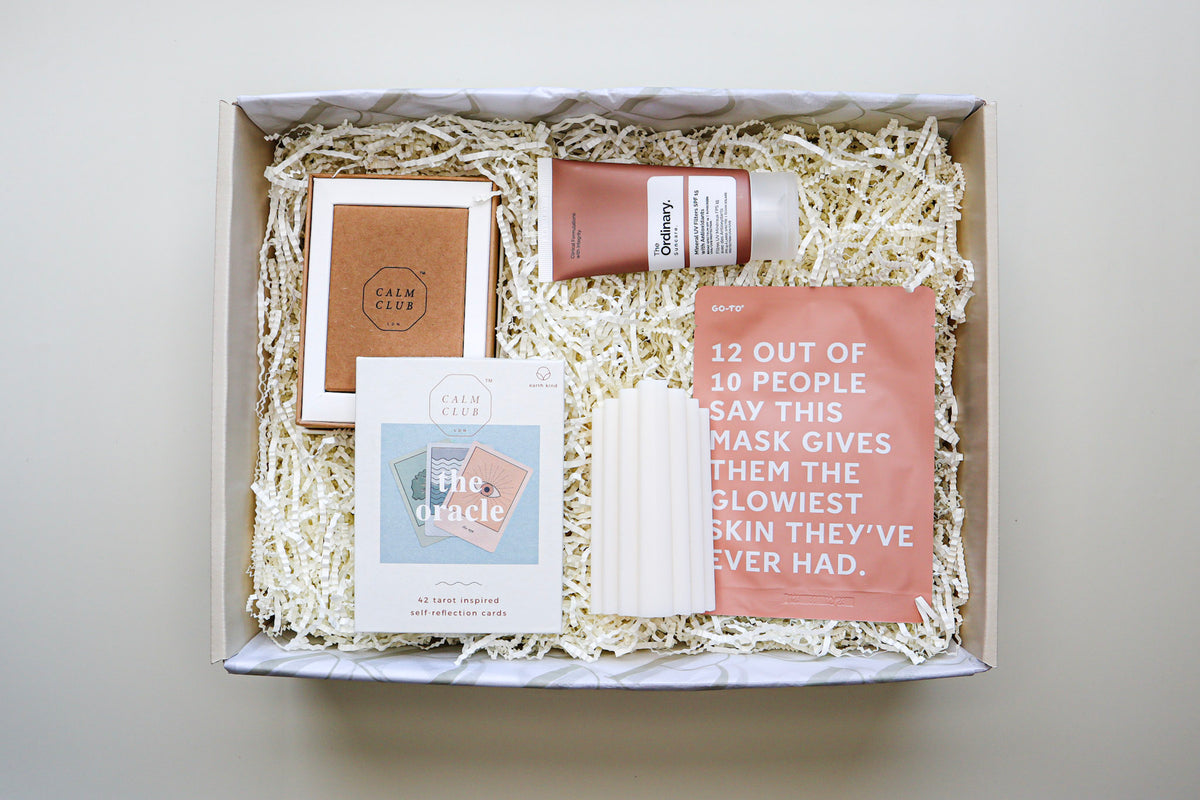 A beige gift box containing The Oracle self-reflection cards in a white box, a white, ridged pillar candle, a tan coloured tube of The Ordinary Sunscreen and a rust coloured Go To Sheet Face Mask