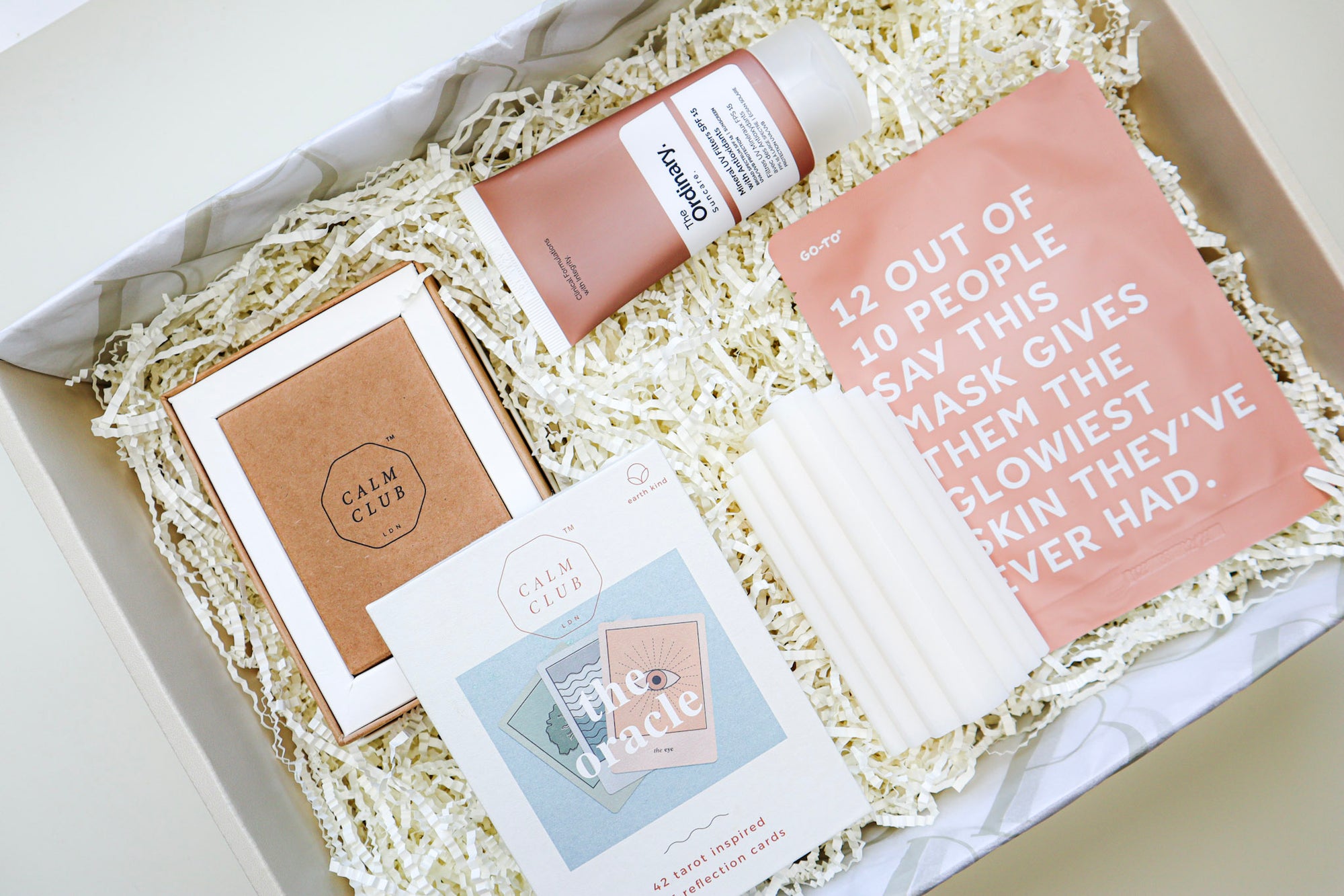 A beige gift box containing The Oracle self-reflection cards in a white and brown cardboard box, a white, ridged pillar candle, a tan coloured tube of The Ordinary Sunscreen and a rust coloured Go To Sheet Face Mask