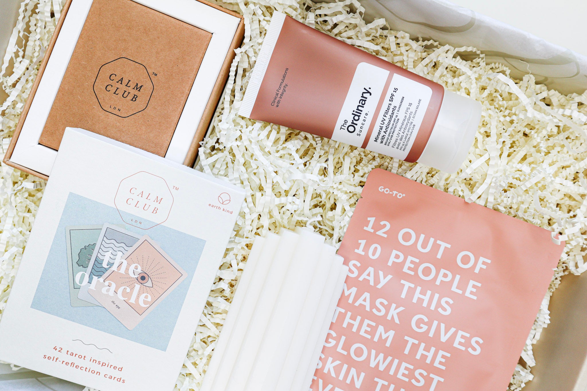 A beige gift box containing The Oracle self-reflection cards in a white and brown cardboard box, a white, ridged pillar candle, a tan coloured tube of The Ordinary Sunscreen and a rust coloured Go To Sheet Face Mask