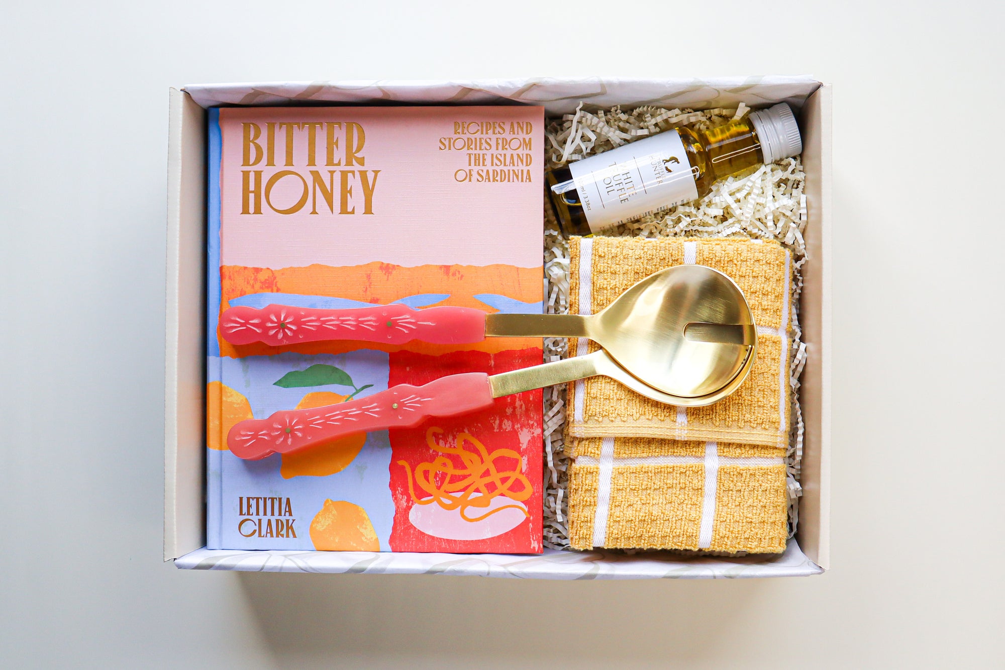 A beige coloured gift box containing a bright hard cover cook book titled Bitter Honey, Gold and Pink Salad Servers, a small bottle of White Truffle Oil and a yellow and white striped tea towel