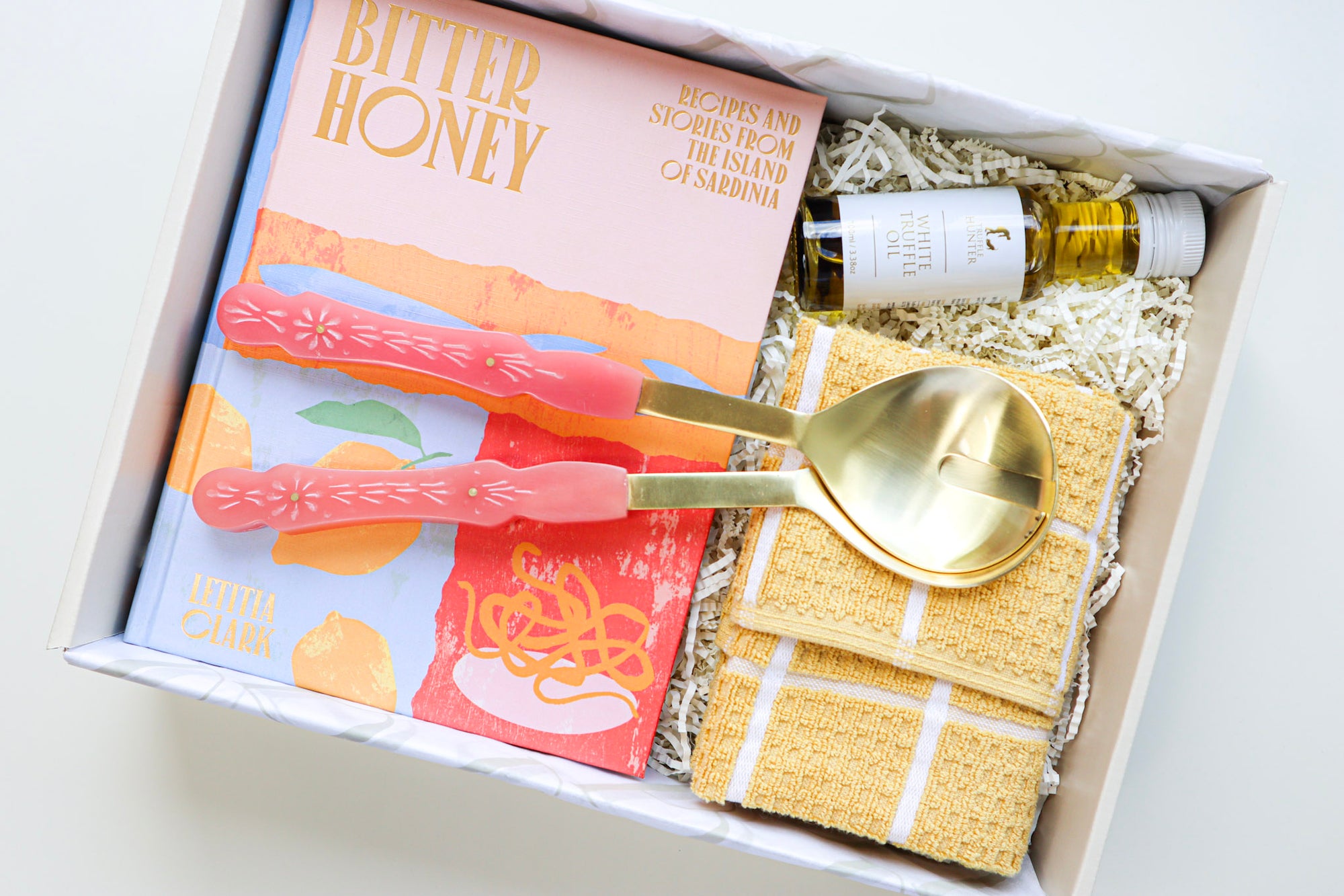 A beige coloured gift box containing a bright hard cover cook book titled Bitter Honey, Gold and Pink Salad Servers, a small bottle of White Truffle Oil and a yellow and white striped tea towel