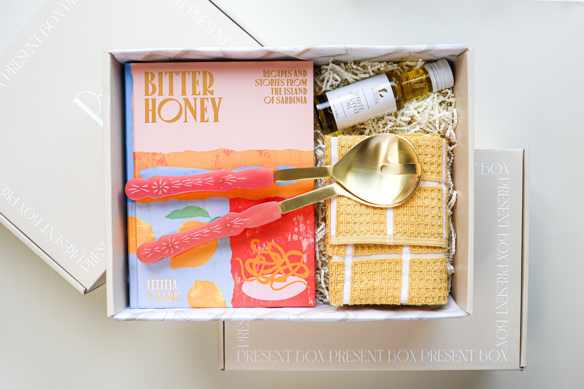A beige coloured gift box containing a bright hard cover cook book titled Bitter Honey, Gold and Pink Salad Servers, a small bottle of White Truffle Oil and a yellow and white striped tea towel