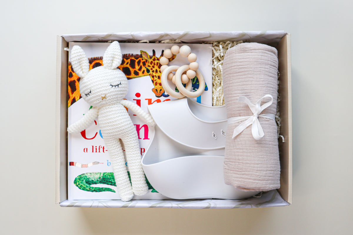 New Baby Gift Box is a cream cardboard box containing a white crocheted bunny toy, a light grey silicone baby bib, a beige cotton wrap rolled and tied with a white ribbon, a white and natural coloured teether ring and rattle toy and a children's hardcover book titled Animal Counting
