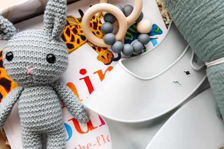 A cute blue, crocheted bunny with black bead eyes and a pink cross-stitched nose, a folded light blue silicone bib, a blue and natural coloured teether ring and rattle toy all lying on top of a children's hardcover book titled Animal Counting