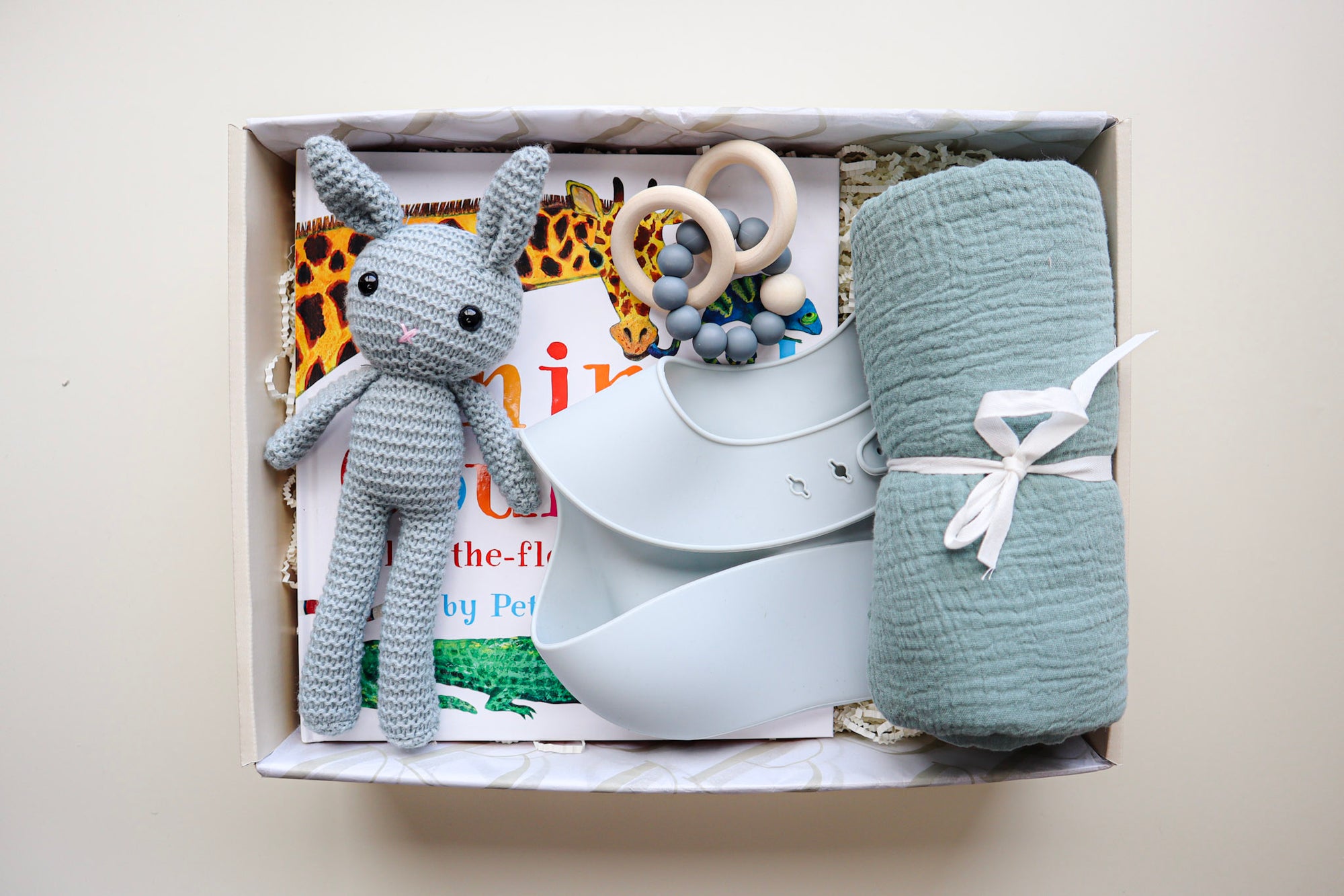 It's A Boy Gift Box is a cream cardboard box containing a blue, crocheted bunny, a light blue silicone bib, a blue cotton wrap rolled and tied with a white ribbon, a blue and natural coloured teether ring and rattle toy and a children's hardcover book titled Animal Counting