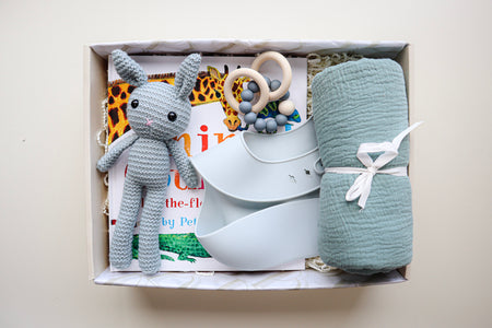 It's A Boy Gift Box is a cream cardboard box containing a blue, crocheted bunny, a light blue silicone bib, a blue cotton wrap rolled and tied with a white ribbon, a blue and natural coloured teether ring and rattle toy and a children's hardcover book titled Animal Counting