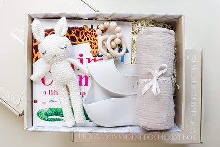 New Baby Gift Box is a cream cardboard box containing a white crocheted bunny toy, a light grey silicone baby bib, a beige cotton wrap rolled and tied with a white ribbon, a white and natural coloured teether ring and rattle toy and a children's hardcover book titled Animal Counting