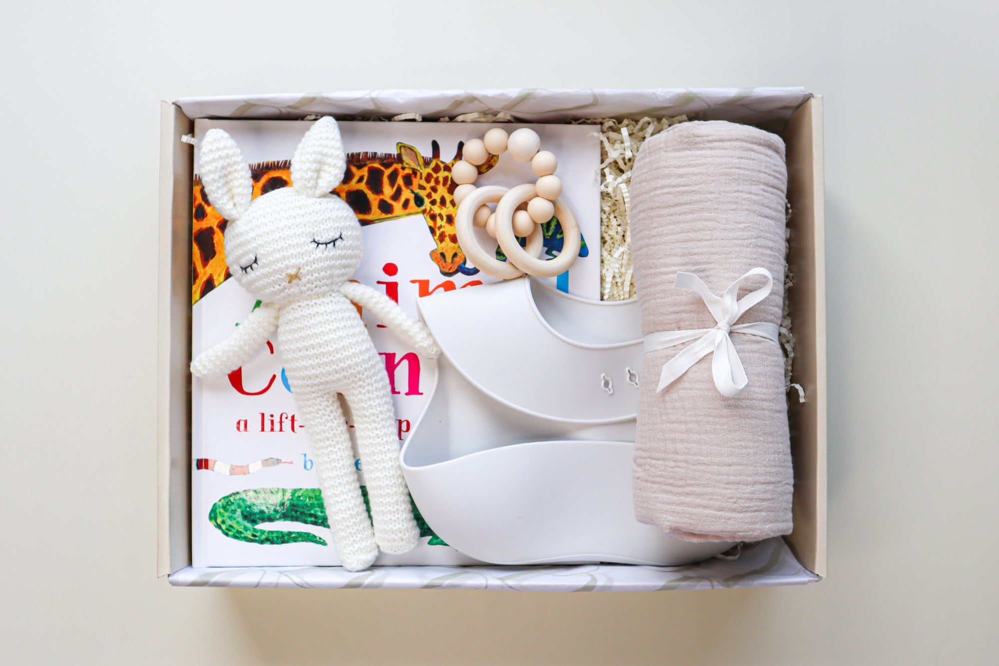 New Baby Gift Box is a cream cardboard box containing a white crocheted bunny toy, a light grey silicone baby bib, a beige cotton wrap rolled and tied with a white ribbon, a white and natural coloured teether ring and rattle toy and a children's hardcover book titled Animal Counting